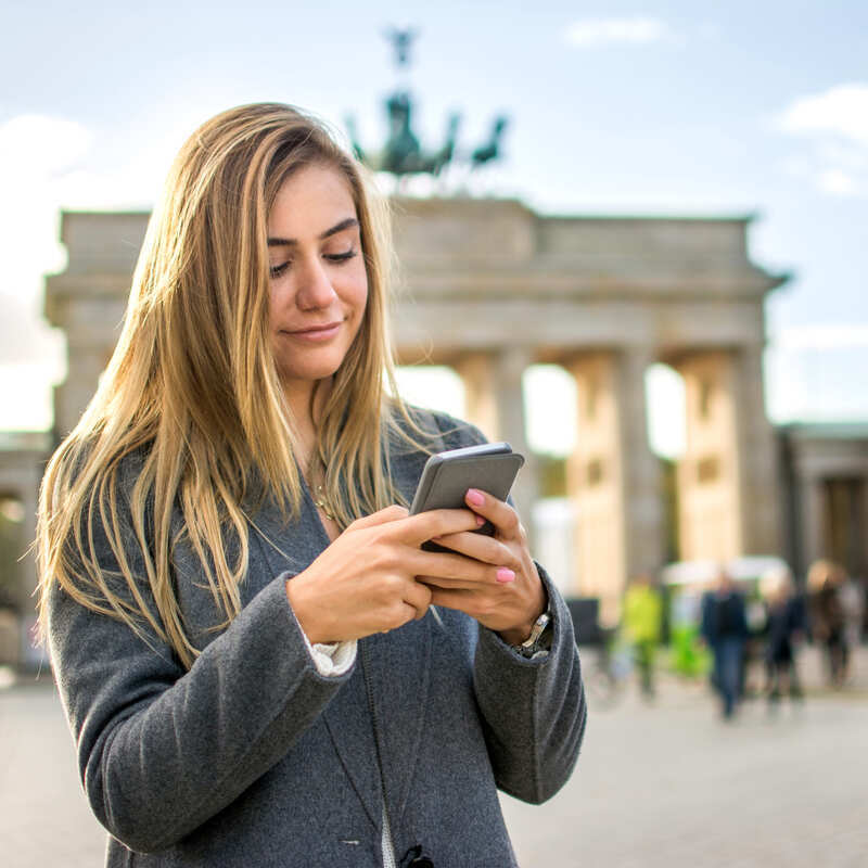 Young Female Tourist Using Her Phone With The Brandenburg Gate In Berlin, Germany For Backdrop
